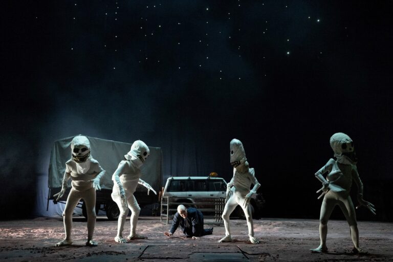 Four human sized Skullie puppets stand on a dark stage and a bald man in a suit crawls on the ground