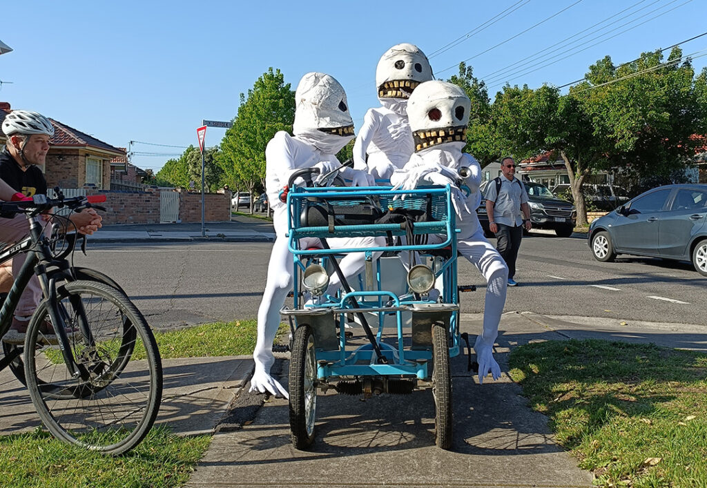 Three Skullie puppets sit on a four seater bike parked on a footpath