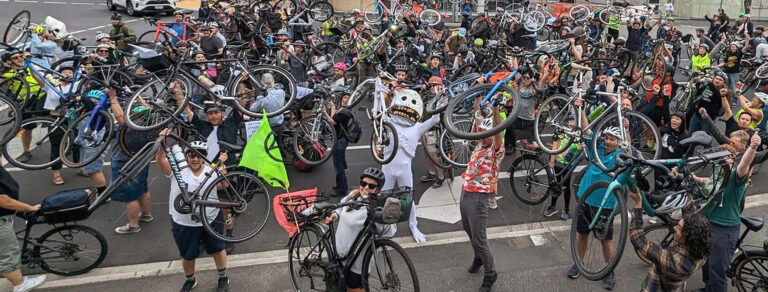 Skullie puppet holds a bike up surrounded by a crowd of people holding bikes up on a road