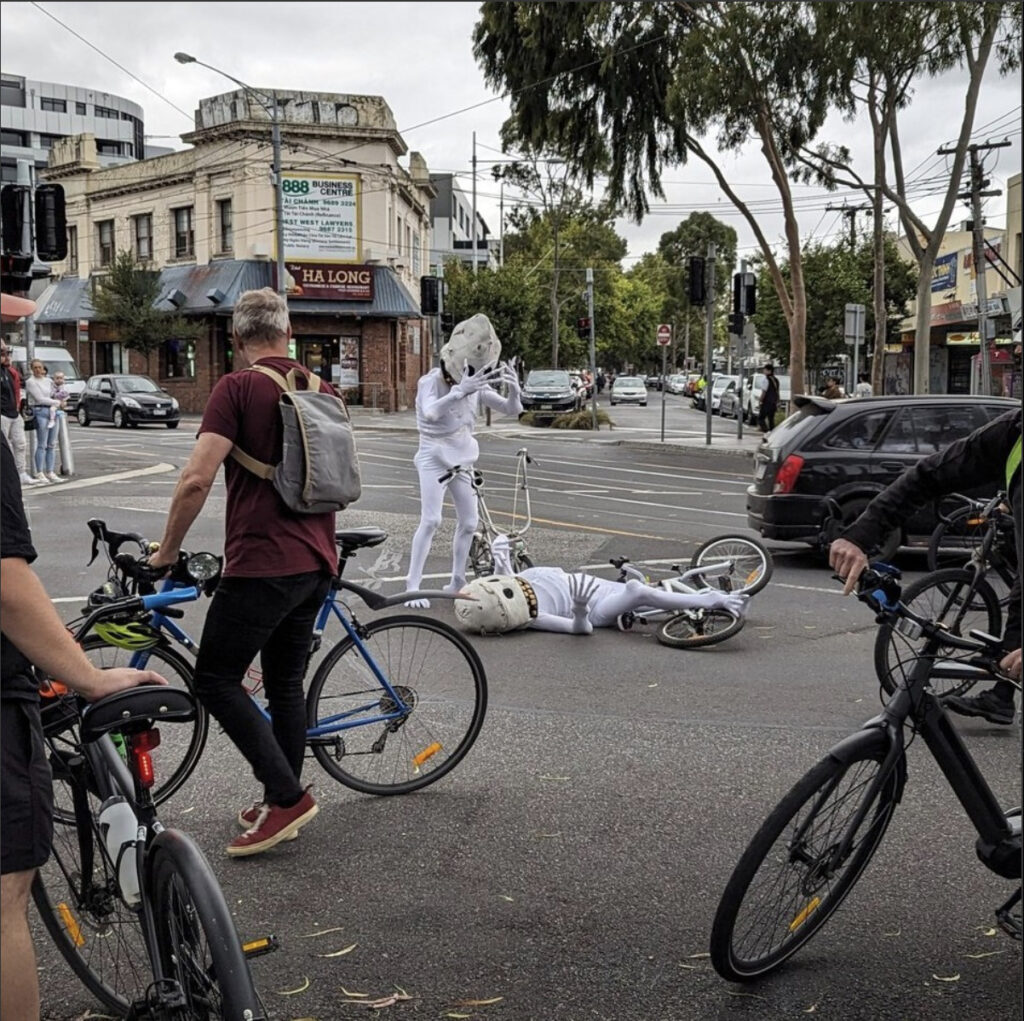 Photo of Barkly St Footscray with a 6-foot tall Skullie puppet laying under a crashed bike on the ground, another Skullie puppet stands above crying, and four bikes and a few cars surround the puppets.